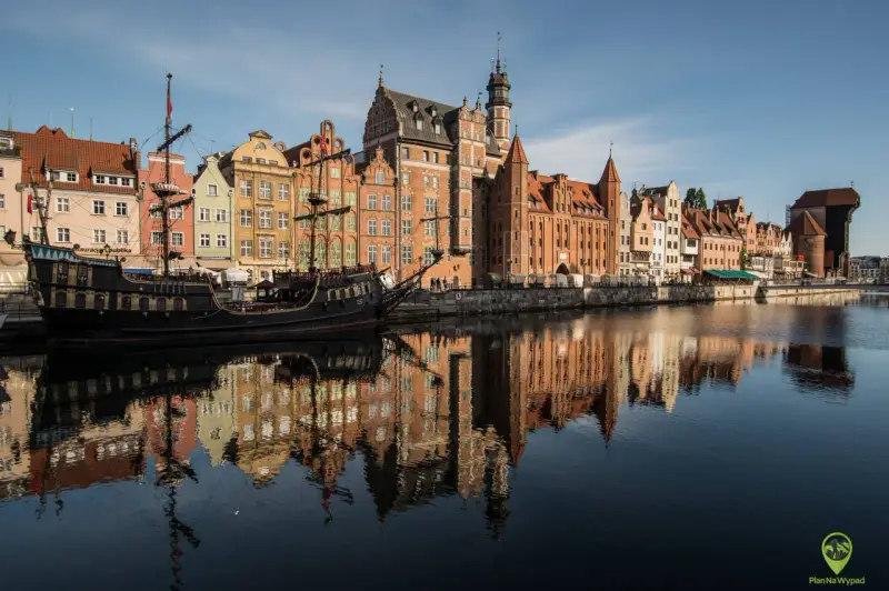 Gdańsk atrakcje: zabytkowy żaglowiec cumuje przy Motławie, w tle kolorowe kamienice i Żuraw. Odbicia budynków w wodzie.